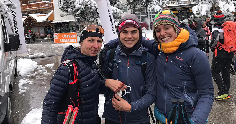 Drie mensen in winterkleding en met rugzakken glimlachen buiten in een besneeuwde omgeving, met op de achtergrond andere mensen en met sneeuw bedekte bomen en gebouwen.